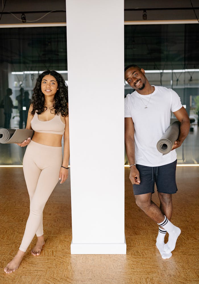 A black man and woman standing indoors with yoga mats, ready for a fitness class.