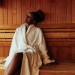 A young black woman is sitting on the bench in the sauna wearing her bathrobe.