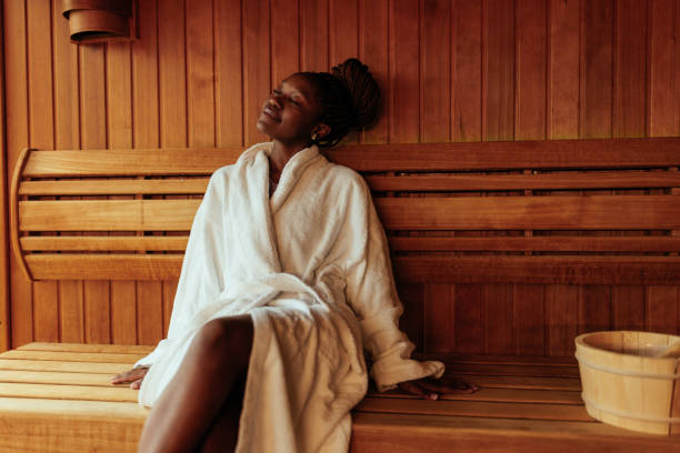 A young black woman is sitting on the bench in the sauna wearing her bathrobe.