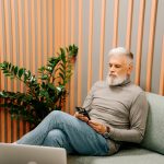 Bearded elderly man with gray sweater texting on smartphone in trendy office lounge.