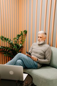 Bearded elderly man with gray sweater texting on smartphone in trendy office lounge.